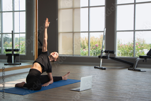 beauty caucasion white woman practicing yoga near window in yoga studio. Young beautiful girl wearing sportswear doing exercises at home with notebook laptop on floor.