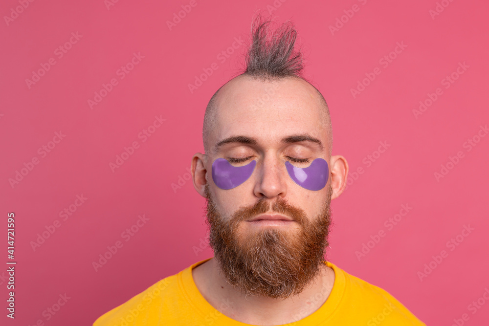 European handsome bearded man in yellow t-shirt and purple eye patches mask, face care for men. Isolated on pink background. European handsome bearded man in yellow t-shirt and purple eye patches mask, face care for men. Isolated on pink background.