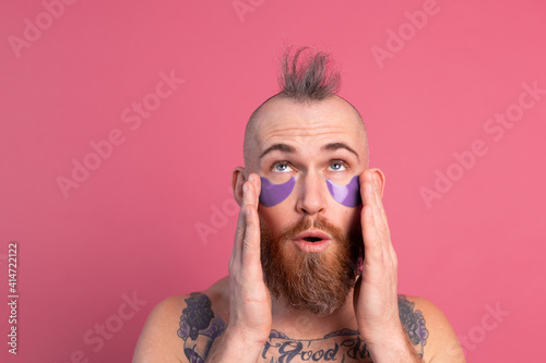European handsome bearded tattooed topless man with purple eye patches mask posing to camera, face care for men. Isolated on pink background.