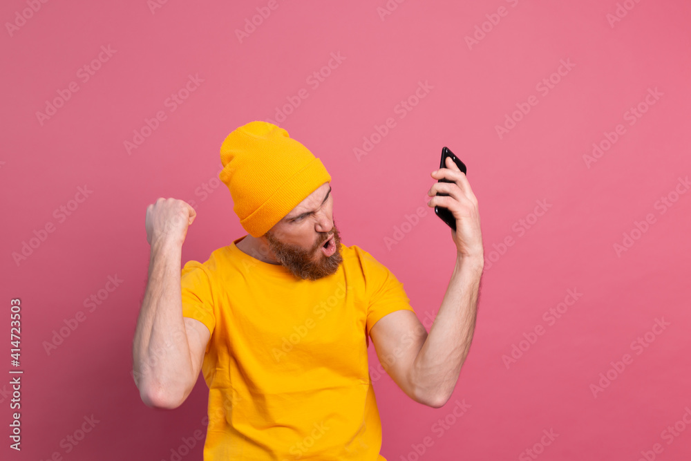 Image of screaming young bearded handsome man standing isolated over pink wall background using mobile phone make winner gesture Image of screaming young bearded handsome man standing isolated over pink wall background using mobile phone make winner gesture
