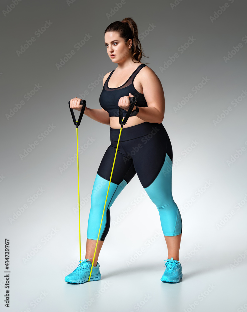 Strong girl working with resistance band. Photo of model with curvy figure in fashionable sportswear on grey background. Sports motivation and healthy lifestyle Strong girl working with resistance band. Photo of model with curvy figure in fashionable sportswear on grey background. Sports motivation and healthy lifestyle
