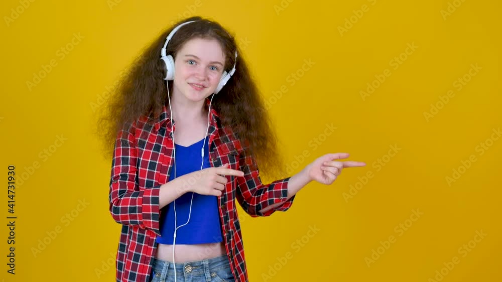 Young funny girl с наушниками look camera pointing fingers hands aside on copy space workspace area isolated on yellow background studio. Young funny girl с наушниками look camera pointing fingers hands aside on copy space workspace area isolated on yellow background studio.