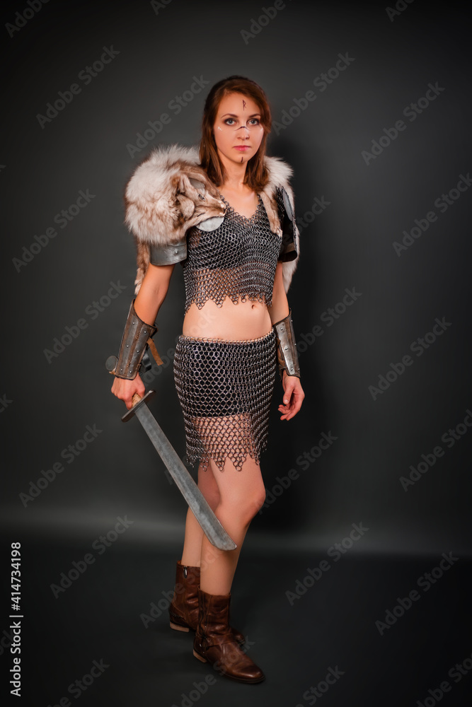 Medieval woman warrior in chain mail armor with lamellar bracers and lamellar shoulder pads with polar fox fur on her shoulders stands with a sword in her hands against a dark background. Medieval woman warrior in chain mail armor with lamellar bracers and lamellar shoulder pads with polar fox fur on her shoulders stands with a sword in her hands against a dark background.