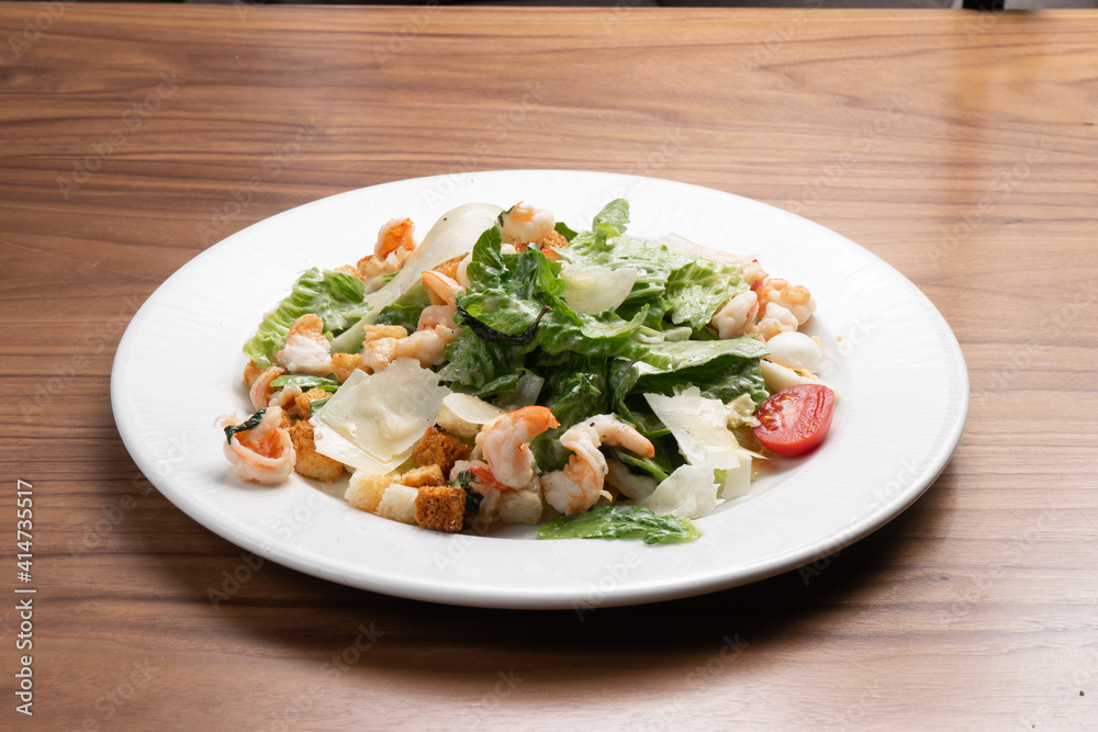 Caesar salad with prawns, lettuce, quail eggs, cherry tomatoes, croutons and parmesan cheese in a white ceramic plate on the wooden kitchen table. Caesar salad with prawns, lettuce, quail eggs, cherry tomatoes, croutons and parmesan cheese in a white ceramic plate on the wooden kitchen table.