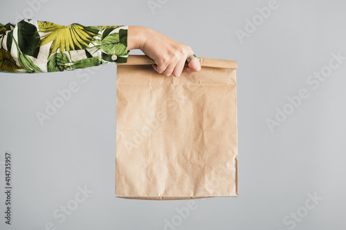 Female hand holds a paper bag against gray backdrop. Empty eco friendly package, gift, surprise or delivery concept photo