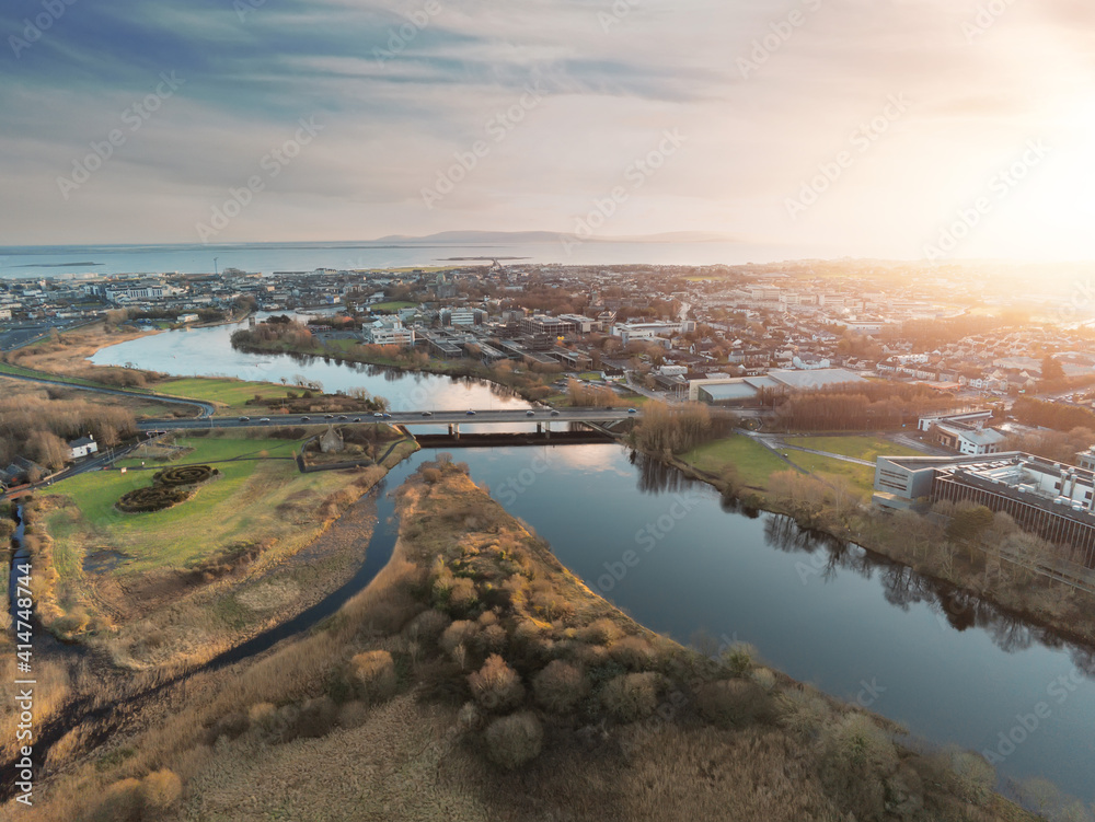 Aerial drone view on Sunset over Galway city, Bridge over River Corrib and NUI buildings, Atlantic ocean in the background. West coast of Ireland. Warm sunny cloudy sky, Sun flare. Warm and cool tone