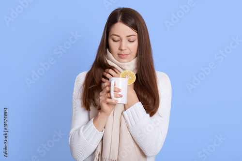 Woman warming up with cup of tea, looking down, keeping hand on neck, has sore throat, wrapped in scarf, wearing white scarf, lady with pleasant appearance enjoying hot beverage against blue wall.