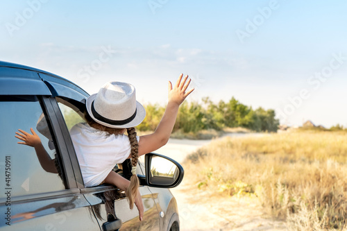 little happy girl in hat raise her arm out of SUV car window with green nature for background. Travel car in vacation.