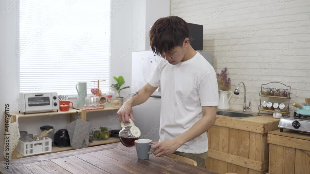 Asian Korean man in casual wear is approaching to the dining table and looking outside while sipping the coffee he just poured from the pot. Asian Korean man in casual wear is approaching to the dining table and looking outside while sipping the coffee he just poured from the pot.