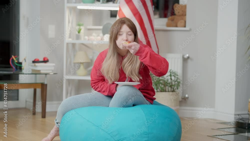 Relaxed Korean teen girl watching TV and eating pizza sitting on bag chair indoors. Wide shot portrait of Asian teenager enjoying leisure at home with American flag at background. Adolescence. Relaxed Korean teen girl watching TV and eating pizza sitting on bag chair indoors. Wide shot portrait of Asian teenager enjoying leisure at home with American flag at background. Adolescence.