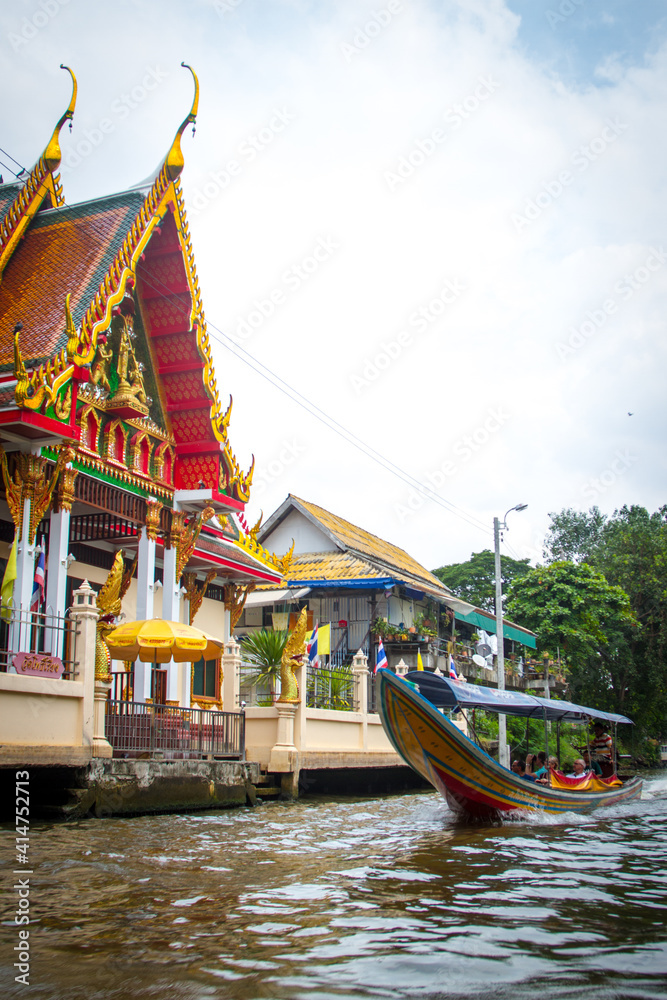 Long tail boat motoring past a buddhist temple in the canals in Bankok, Thailand. 