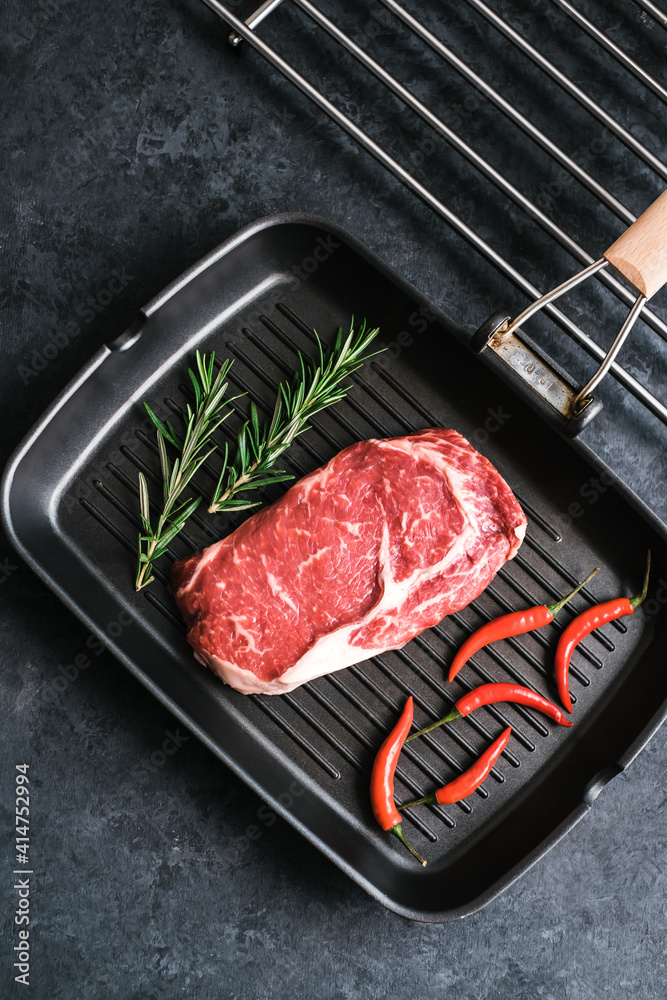Raw rib eye beef steak on black grilling pan with chili pepper and rosemary, top view.