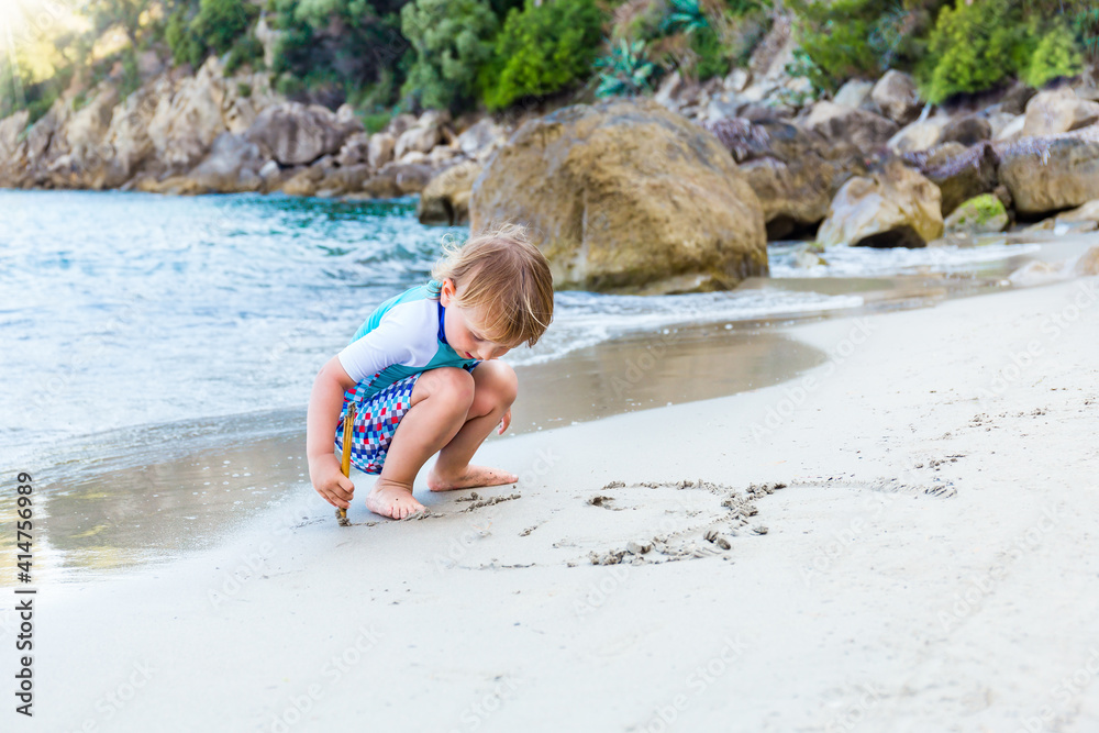 Adorable blond toddler boy plays on the beach at the seaside. A young boy drawing with bamboo stick on the sand near the sea during summer holidays. Adorable blond toddler boy plays on the beach at the seaside. A young boy drawing with bamboo stick on the sand near the sea during summer holidays.