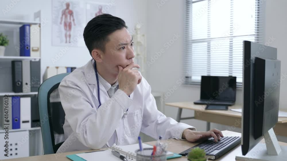 portrait asian physician is touching his chin in contemplation while looking at his note and continues typing on the desktop in the office.