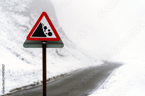 Warning sign red triangle falling rocks on icy snow covered winding mountain road passing through foggy and mist filled valley between hills during winter season
