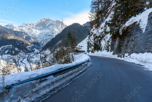 Winding alpine road in a snowy forested mountain landscape on a sunny winter day. A small town along the banks of a river running at the bottom of a valley is visible on the left side of the picture. © alpegor