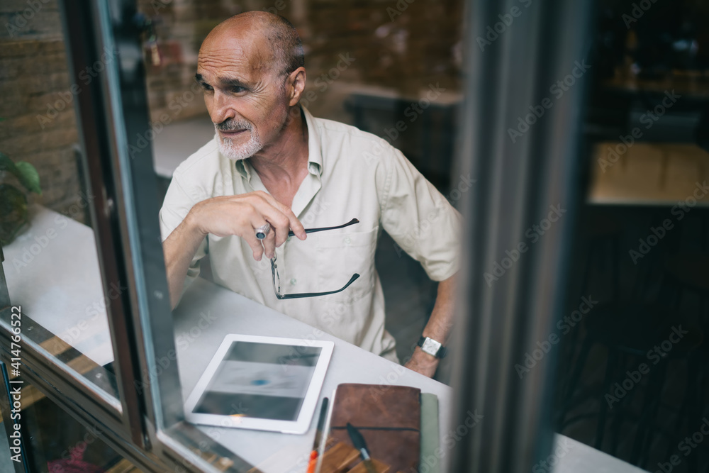 Fototapeta premium Window view of old Caucaisan journalist with optical glasses in hand thoughtful looking away, elderly male blogger with modern touch pad for browsing thinking about web information in cafe interior
