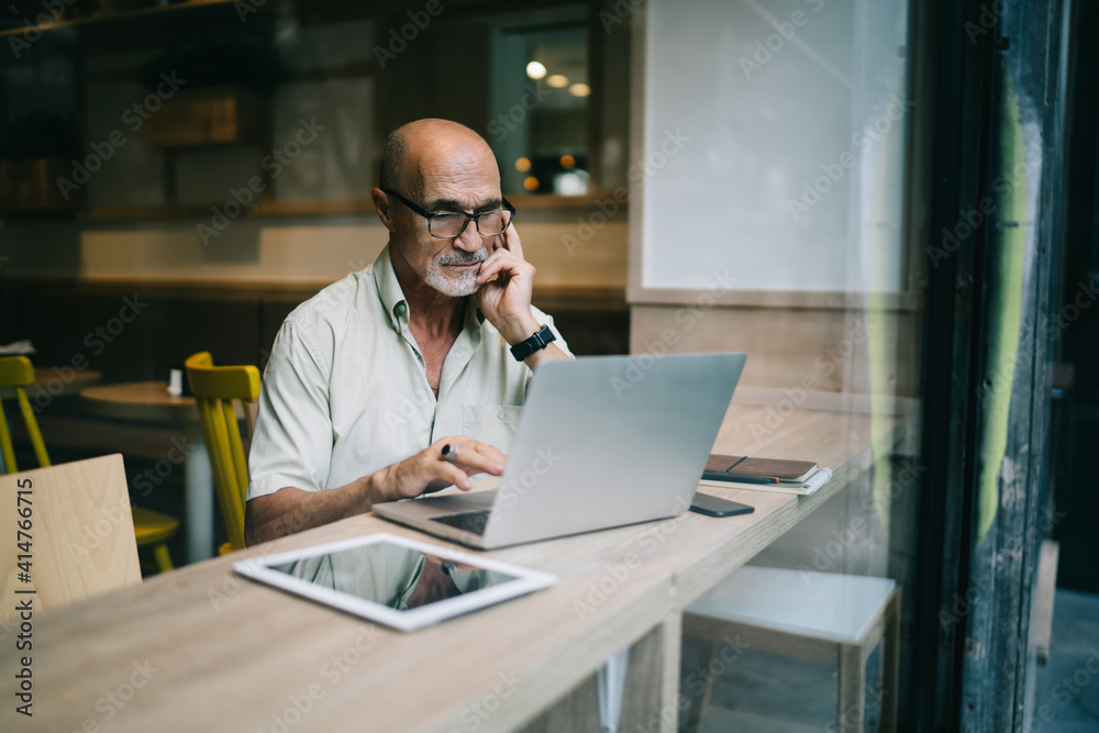 Mature man 60s sitting in coworking cafeteria with modern netbook and working remotely with project, aged Caucasian male in optical eyewear watching online video during laptop banking at table desktop