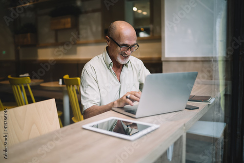 Mature cheerful man in spectacles typing on laptop computer share content in blog browsing web pages and information, happy male freelancer doing remote job using netbook in coworking and smiling