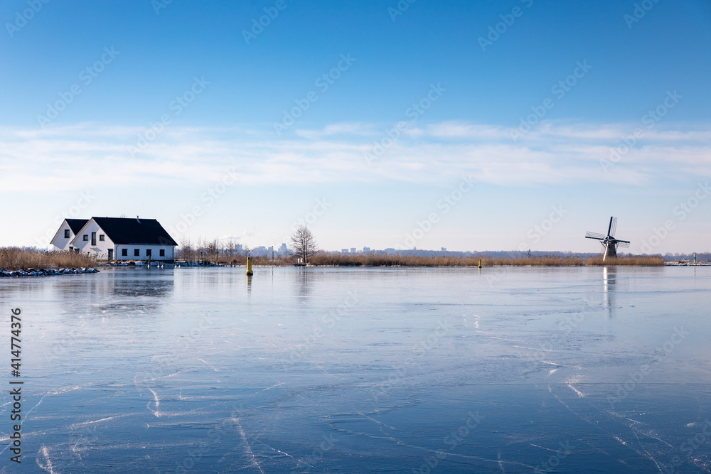 Fototapeta premium Winterlandscape with ice on the Dieperpoel (Kagerplassen) in front of a windmill (de Kok) and house on the Kogjespolder in the south-holland municipality of Teylingen (Sassenheim) in the Netherlands.