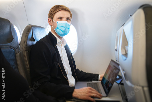 A man wearing face mask while sitting into an airplane. New normal traveling during a pandemic. Male passenger traveling. Wearing FFP2 mask in aircraft cabin. Travel Covid-19 Work from plane on laptop