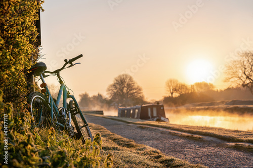 Canal boat with mountain bike left leaning against hedge row early morning sunrise dawn with golden light in sky on the River Trent and mist rising in Nottingham biker on ride out photo
