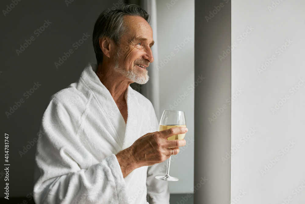 Smiling senior man with wineglass. Side view of a bearded mature male wearing a bathrobe in a hotel room looking at window. Smiling senior man with wineglass. Side view of a bearded mature male wearing a bathrobe in a hotel room looking at window.