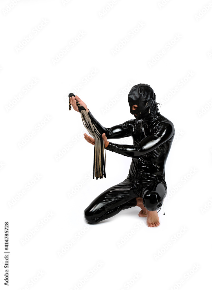 Slave sitting on his knees. Man dressed in latex rubber fetish clothes and pvc mask. Slave bowed his knees and holds a leather whip in hands. bdsm costume for role-playing adult games. Slave sitting on his knees. Man dressed in latex rubber fetish clothes and pvc mask. Slave bowed his knees and holds a leather whip in hands. bdsm costume for role-playing adult games.
