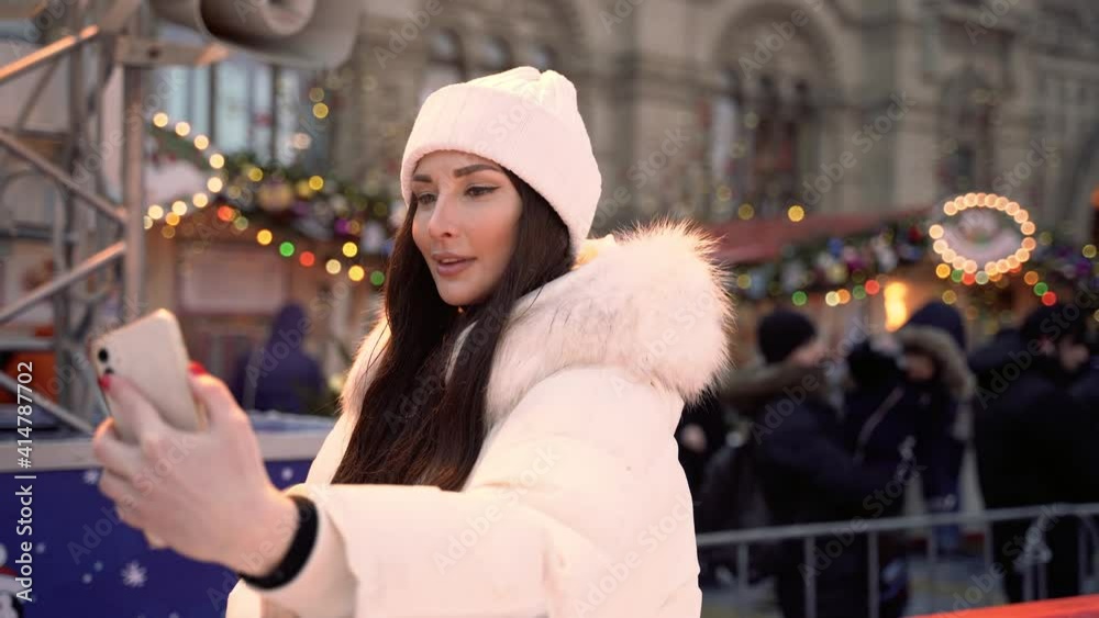 A pretty brunette european girl stands at the side of the ice rink makes a selfie on the phone against the background of the christmas fair High quality 4k footage A pretty brunette european girl stands at the side of the ice rink makes a selfie on the phone against the background of the christmas fair High quality 4k footage