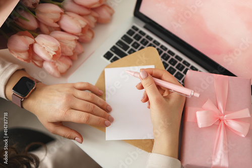 Female hands writing down wishes on latter paper for Valentines or Birthday with gift box and laptop on desk, top view photo