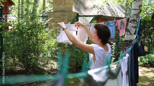 Candid real life portrait of young adult beautiful attractive caucasian woman hanging up fresh washed family clothes on birch tree clothesline with pins at home yard on bright sunny day outdoors