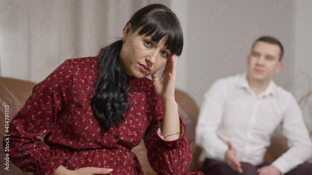 Portrait of sad stressed pregnant woman rubbing temples and stroking belly as blurred man yelling and gesturing at background. Unhappy Caucasian expectant arguing with husband at home.