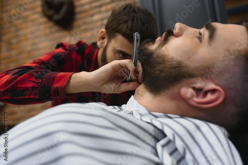 Serious Bearded Man Getting Beard Haircut With A Straight Razor By Barber While Sitting In Chair At Barbershop. Barbershop Theme