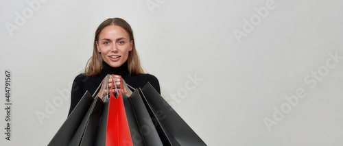 Studio shot of young caucasian woman wearing black clothes looking happy, holding bunch of shopping bags in front of her, posing isolated over light gray background