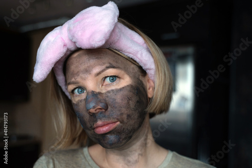 a girl with blue eyes and a cleansing charcoal mask on her face and a headband and turns her head