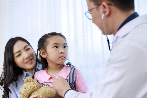 asian doctor in white coat using stethoscope to listen to heart and lung sound of young girl