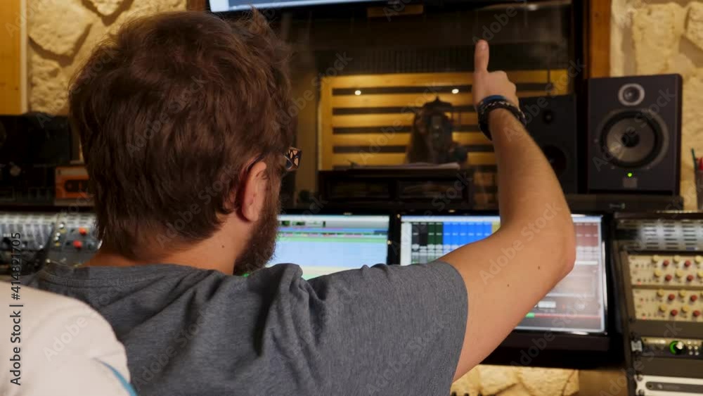 Rear view closeup of a sound producer giving thumbs up while listening to pop artist singing in booth in recording studio Rear view closeup of a sound producer giving thumbs up while listening to pop artist singing in booth in recording studio