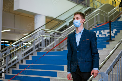 Young caucasian man in a classic jacket wearing a medical mask on the steps in a shopping mall
