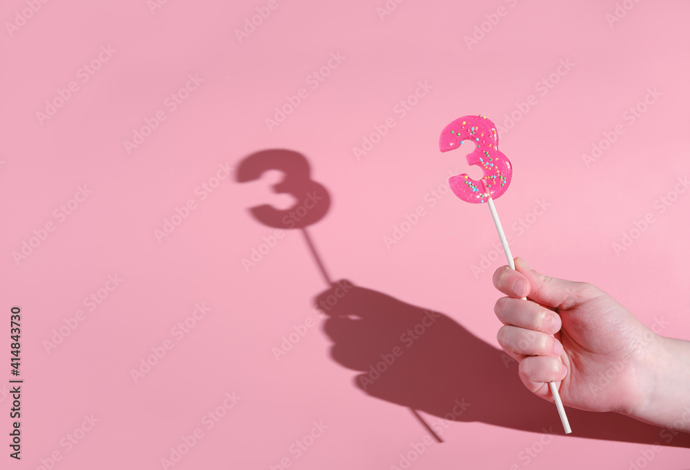 girl's hand holding a number 3 shaped lolipop. anniversary or birthday selebration. kids party sweet treats. pink background and direct light. girl's hand holding a number 3 shaped lolipop. anniversary or birthday selebration. kids party sweet treats. pink background and direct light.
