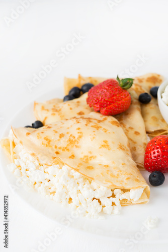 Traditional Russian thin pancakes with cottage cheese and berries in a plate on a white background