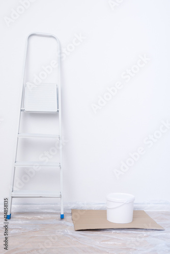 a folding ladder and a white paint bucket stand against a white wall, preparing for indoor finishing work