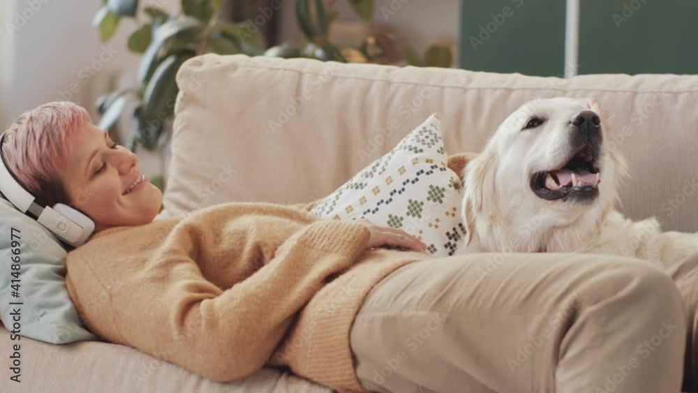 Cheerful woman in wireless headphones lying on sofa at home and petting cute golden retriever dog Cheerful woman in wireless headphones lying on sofa at home and petting cute golden retriever dog