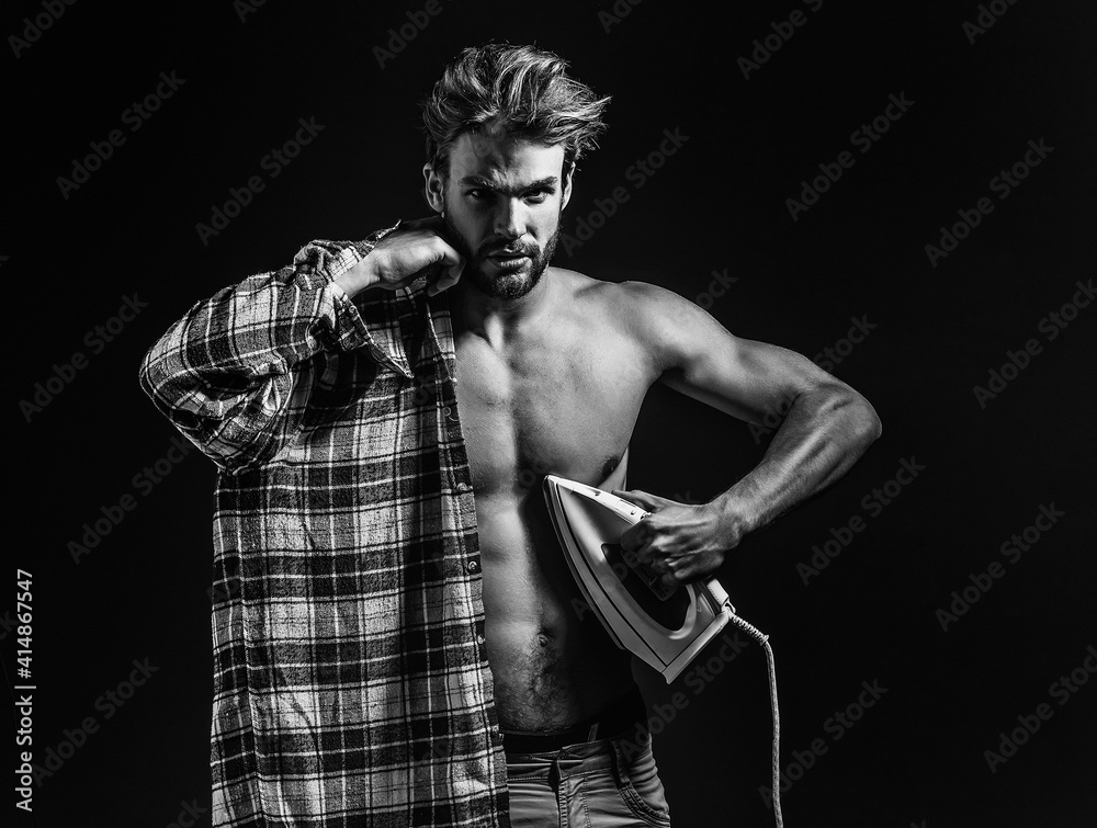 Sexy young shirtless man with messy hair, irons plaid shirt on black background.