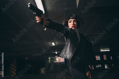 Beautiful brunette sexy spy agent (killer or police) woman in leather jacket and jeans with a gun in her hand running after someone, to catch him on parking