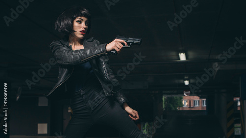 Beautiful brunette sexy spy agent (killer or police) woman in leather jacket and jeans with a gun in her hand running after someone, to catch him on parking