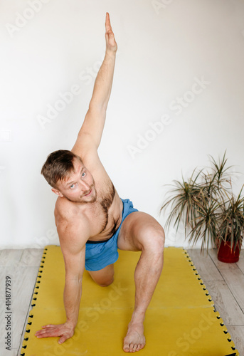 a man of European appearance performs a stretching exercise in a bright room at home. An athlete with a naked torso stands on one knee, raises his hand up, looks at the camera