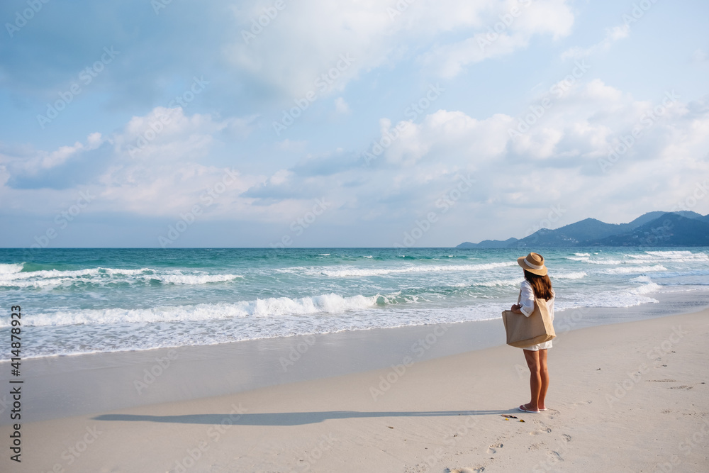 Rear view image of a woman with hat and bag strolling on the beach with blue sky background