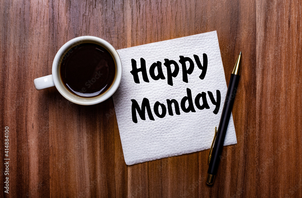 On a wooden table next to a white cup of coffee and a pen is a white paper napkin with the words HAPPY MONDAY On a wooden table next to a white cup of coffee and a pen is a white paper napkin with the words HAPPY MONDAY