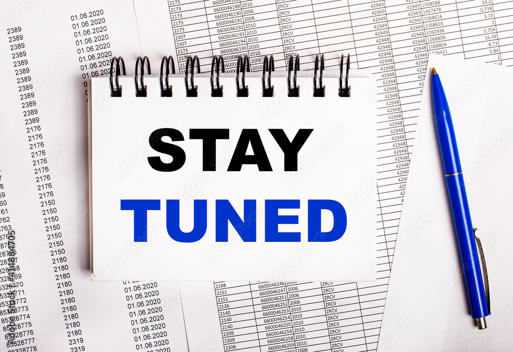 On the table are charts and reports, on which lie a blue pen and a notebook with the words STAY TUNED On the table are charts and reports, on which lie a blue pen and a notebook with the words STAY TUNED