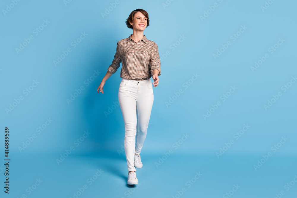 Full size portrait of confident cheerful lady walking look empty space beaming smile isolated on blue color background Full size portrait of confident cheerful lady walking look empty space beaming smile isolated on blue color background